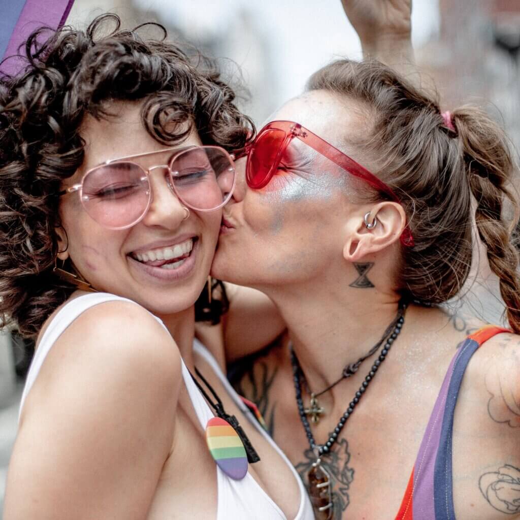 Portrait of two individuals outside at pride parade wearing colorful clothing and sunglasses embracing