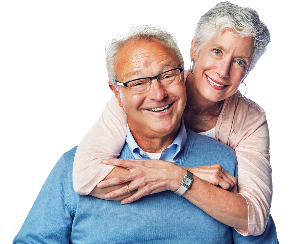 An elderly couple, both smiling, pose affectionately. The man, in a blue sweater and glasses, stands while the woman, in a pink sweater, embraces him from behind. They both have short grey hair and radiate health. The background is white.