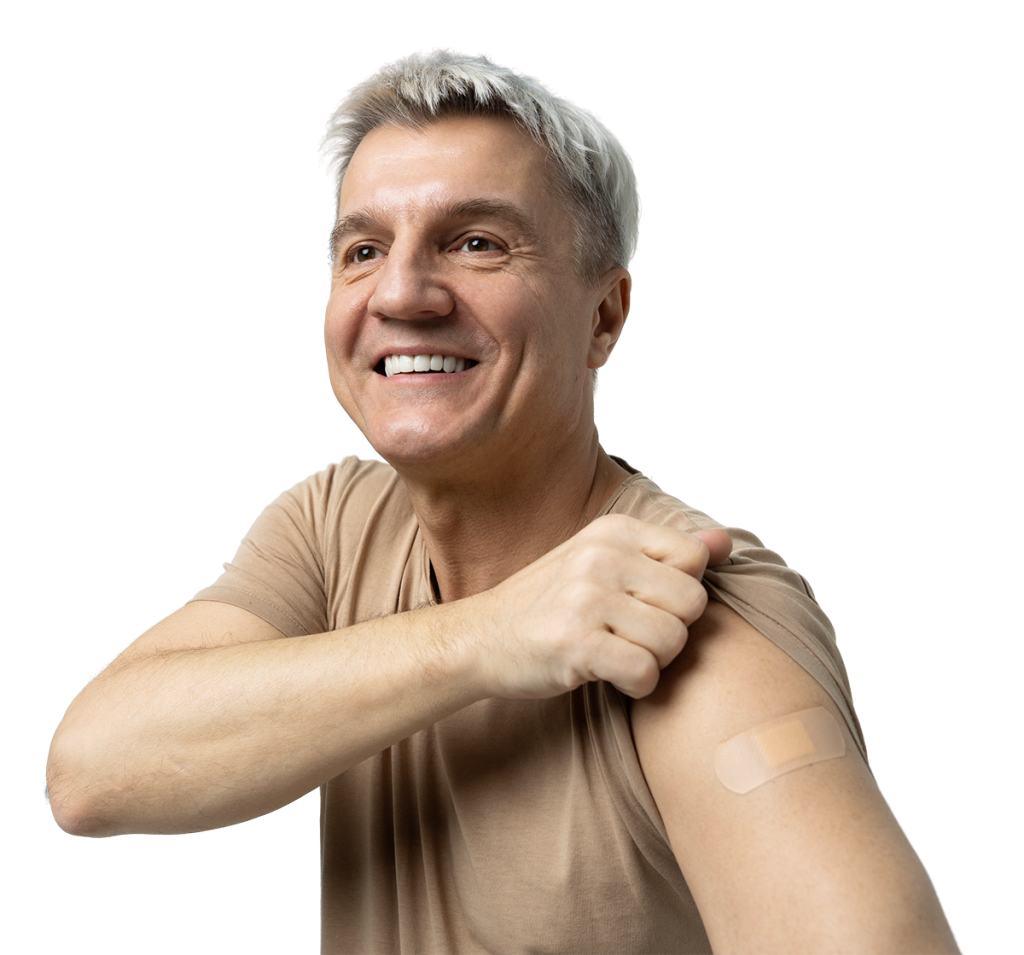 A smiling older adult with short gray hair and wearing a brown t-shirt proudly points to a bandage on their upper arm, indicating they have received a vaccination or got blood testing. The background is white.