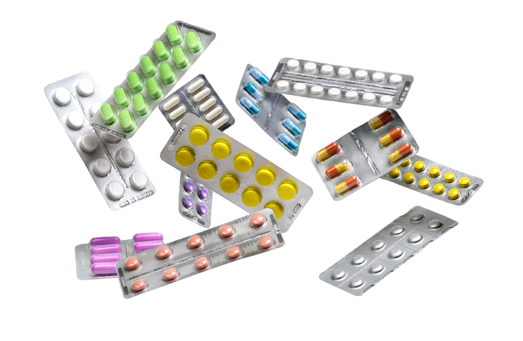 Various blister packs of tablets and capsules in different colors and shapes are scattered in a random arrangement. The packs contain pills in shades of green, purple, yellow, white, blue, red, and orange.