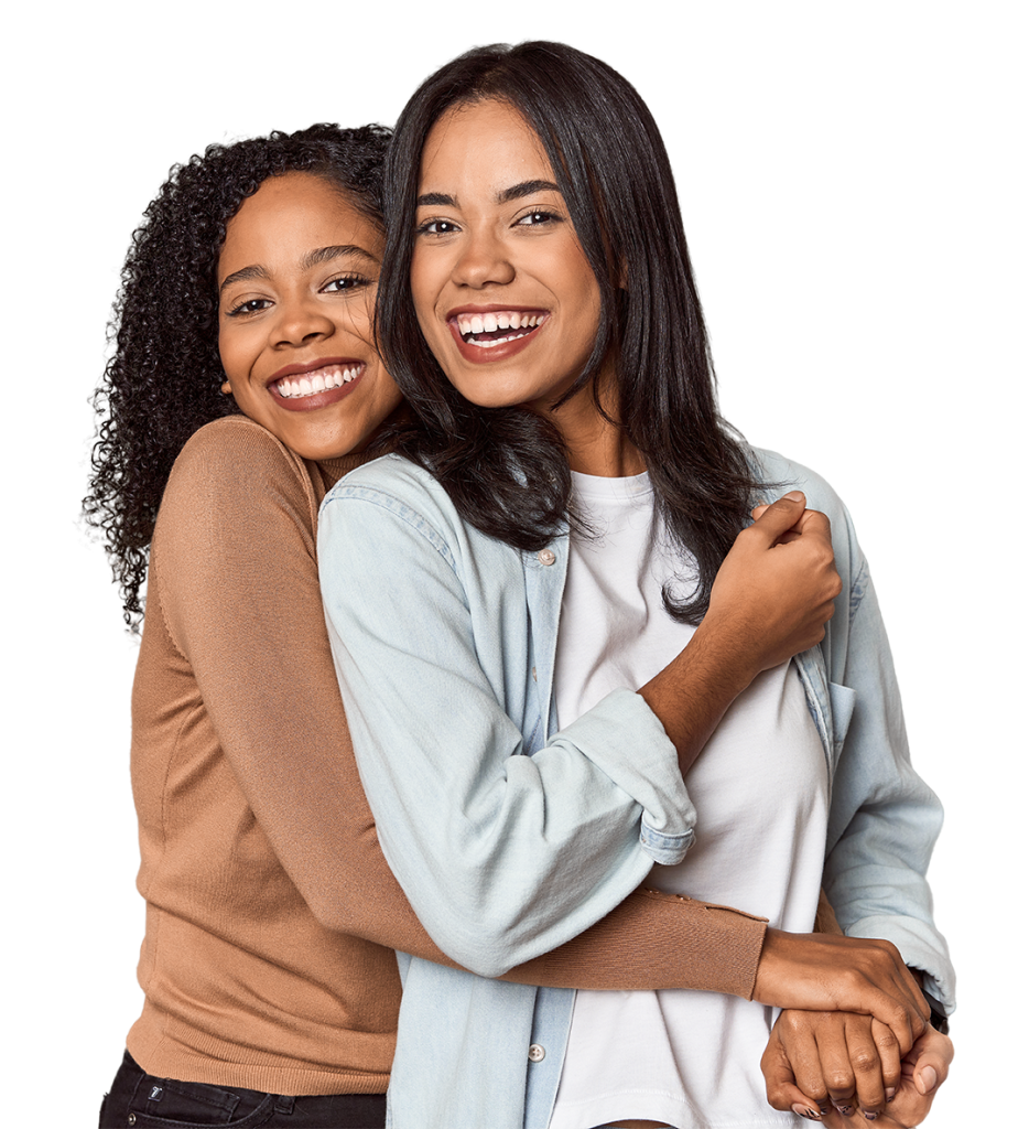 Two women are standing and smiling at the camera. One woman, in a light blue shirt, has her arm around the other woman, who is wearing a brown sweater. Both have long, dark hair. They appear happy and close, embracing each other warmly.