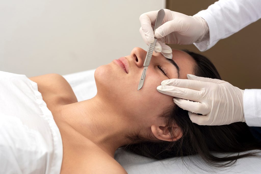 A woman lying on a treatment bed with her eyes closed as a professional wearing white gloves performs a skincare procedure on her face using a scalpel. The woman is covered with a white towel.