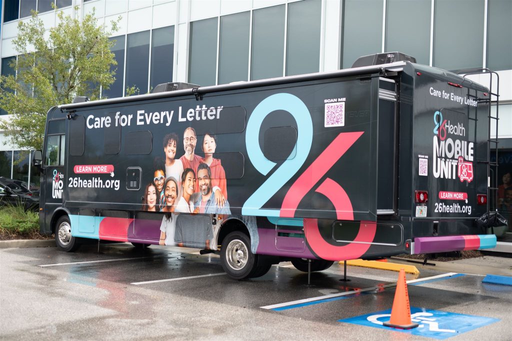 A black mobile health unit van is parked in a lot in front of a building. The van is adorned with the number "26" and images of diverse individuals. It promotes the website "26health.org" with the slogan “Care for Every Letter.” There are colorful designs along the bottom.