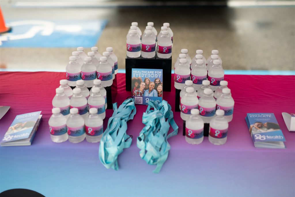 A table with water bottles, brochures, and blue lanyards arranged neatly on a colorful tablecloth. A central sign reads "Healthcare for Everybody" with a picture of diverse individuals. The setup is for a health-related event or promotional activity.