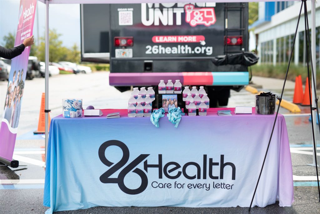 A display table is seen at an outdoor event, with the tablecloth reading "26Health Care for every letter." The table has folded pink pamphlets, bottled water, and small items. Behind the table, a van with the 26Health logo, website, and slogan is visible.