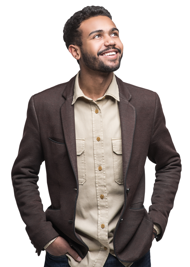 A man with short, dark curly hair and a beard is smiling and looking upwards. He is wearing a brown blazer over a beige button-up shirt with front pockets. His hands are in his pants pockets, and he has a relaxed, confident posture.