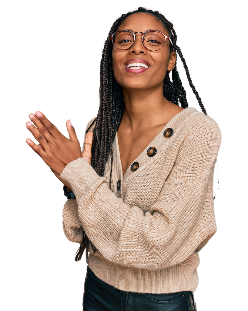 A person with long braided hair and glasses is smiling with their mouth open, wearing a beige sweater with button details and black pants. Their hands are clasped together near their chest.