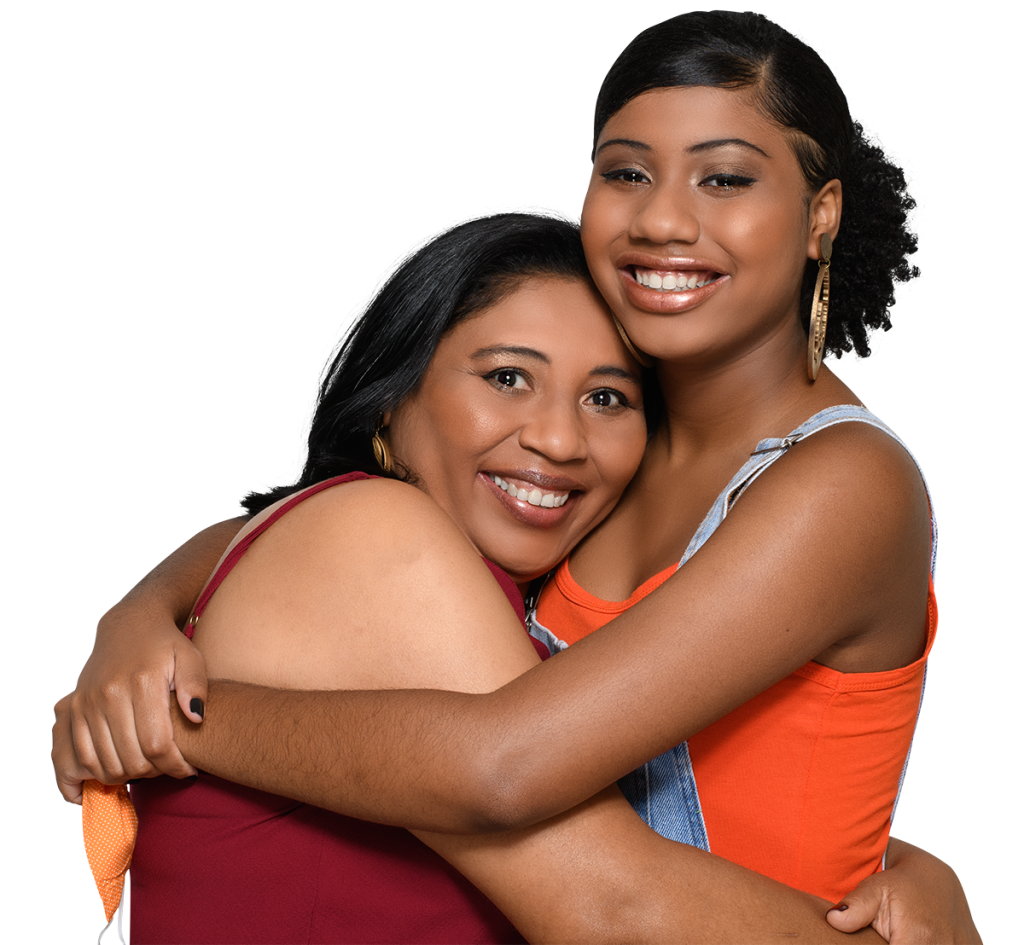 Family and Relationship Therapy. Two women are smiling and hugging each other. The woman on the left has shoulder-length black hair and wears a maroon top. The woman on the right has curly hair pulled back, gold hoop earrings, and wears an orange top with denim overalls. Both look happy and affectionate.