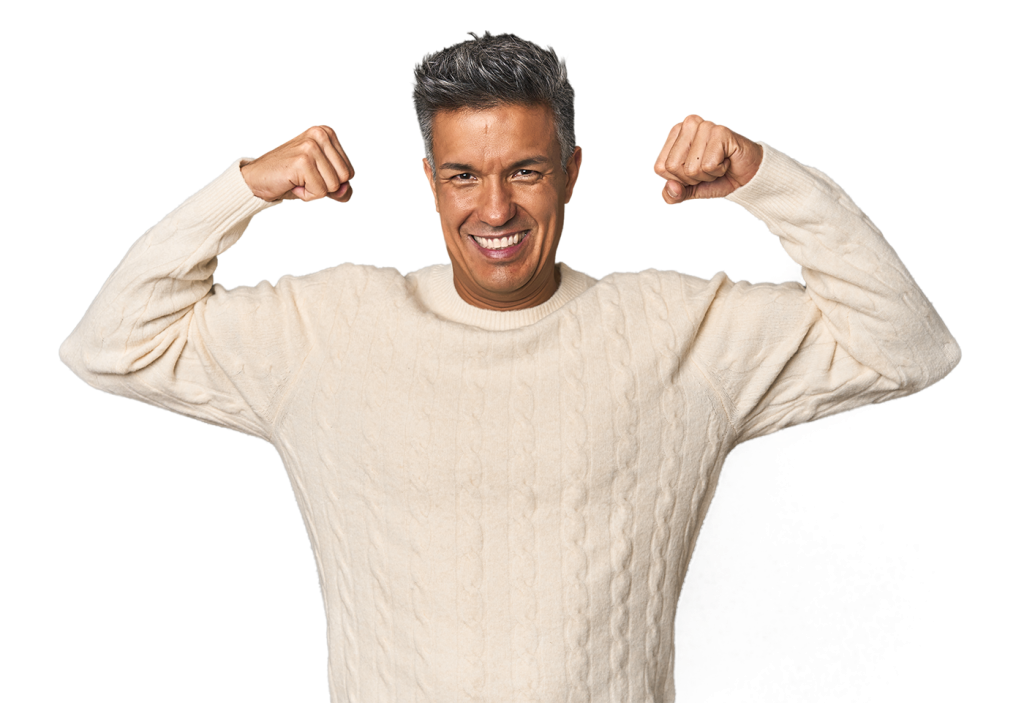 A man in a beige cable-knit sweater is smiling broadly and flexing his arms with both fists raised, displaying a confident and joyful demeanor. The background is white.