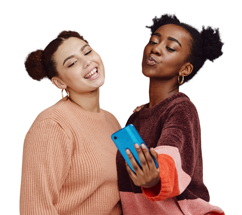 Two women taking a selfie together. One woman with curly hair is smiling while the other woman with curly hair is making a kissing face. Both are wearing cozy sweaters in shades of pink and holding a blue smartphone.